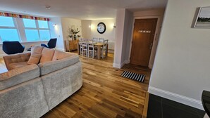 A living room with a grey sofa, wooden flooring, a dining area with a table and chairs, a clock on the wall, and a wooden door.