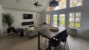 A spacious living area with a high ceiling, a ceiling fan, a large window with a view of the outdoors, a flat-screen TV mounted on the wall, and a dining table with chairs. - Lakefront Cottage with Sauna- #3 Chinook (Wasilla)