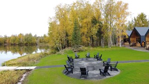 Uma área de estar ao ar livre na beira do lago, com disposição circular de cadeiras e uma lareira no centro, cercada por um gramado e árvores com folhagem outonal.