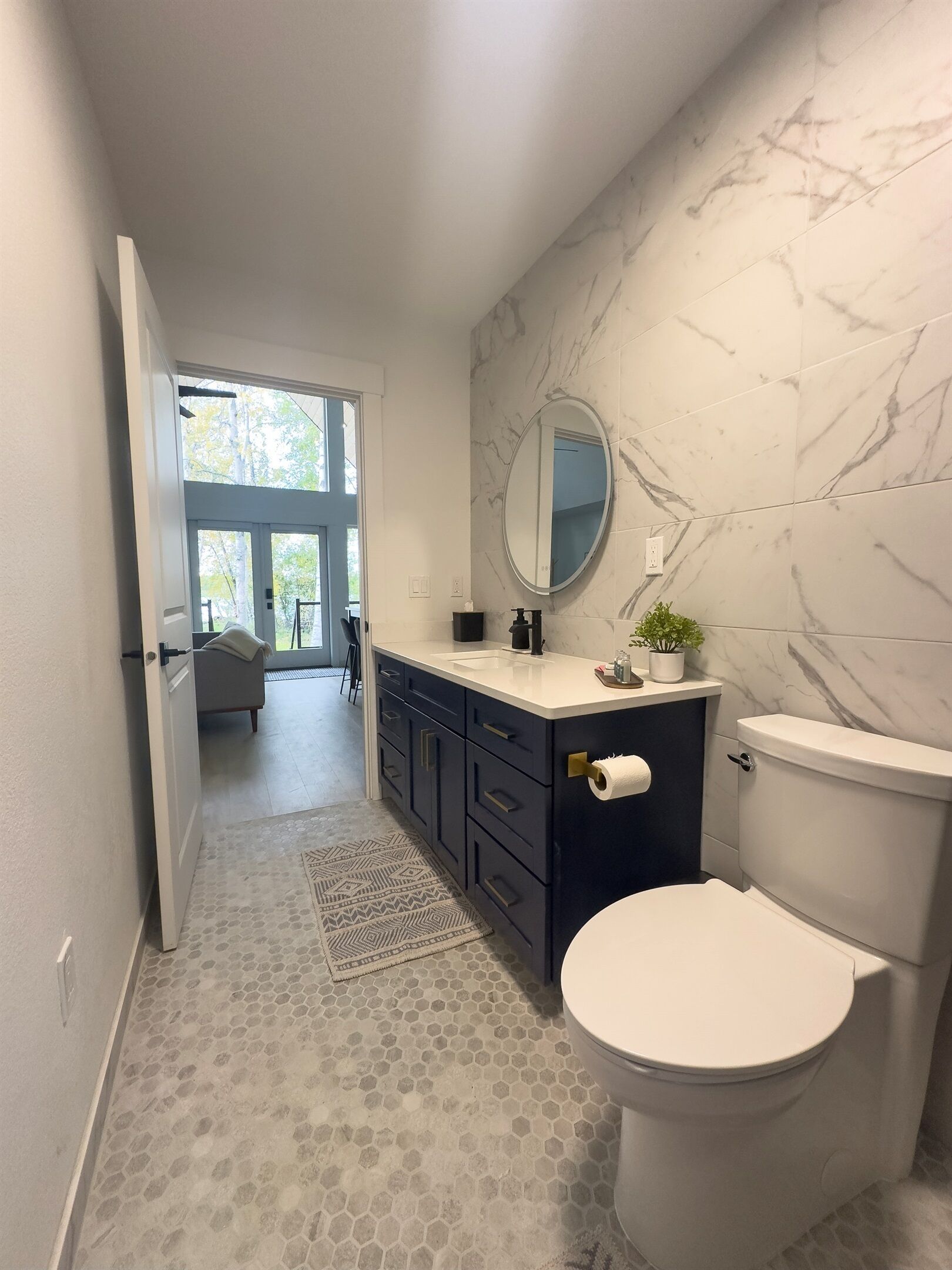 A modern bathroom with a marble backsplash, a round mirror, a toilet, and a sink with a dark cabinet.
