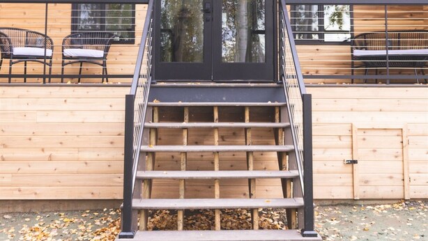 A wooden cabin with a staircase leading to a balcony, large windows, and a view of trees. - Lakefront Cottage with Sauna - #7 Dolly Varden (Wasilla)