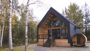 A modern log cabin with a large window, a wooden deck, and a barrel-shaped door. - Lakefront Cottage with Sauna - #7 Dolly Varden (Wasilla)