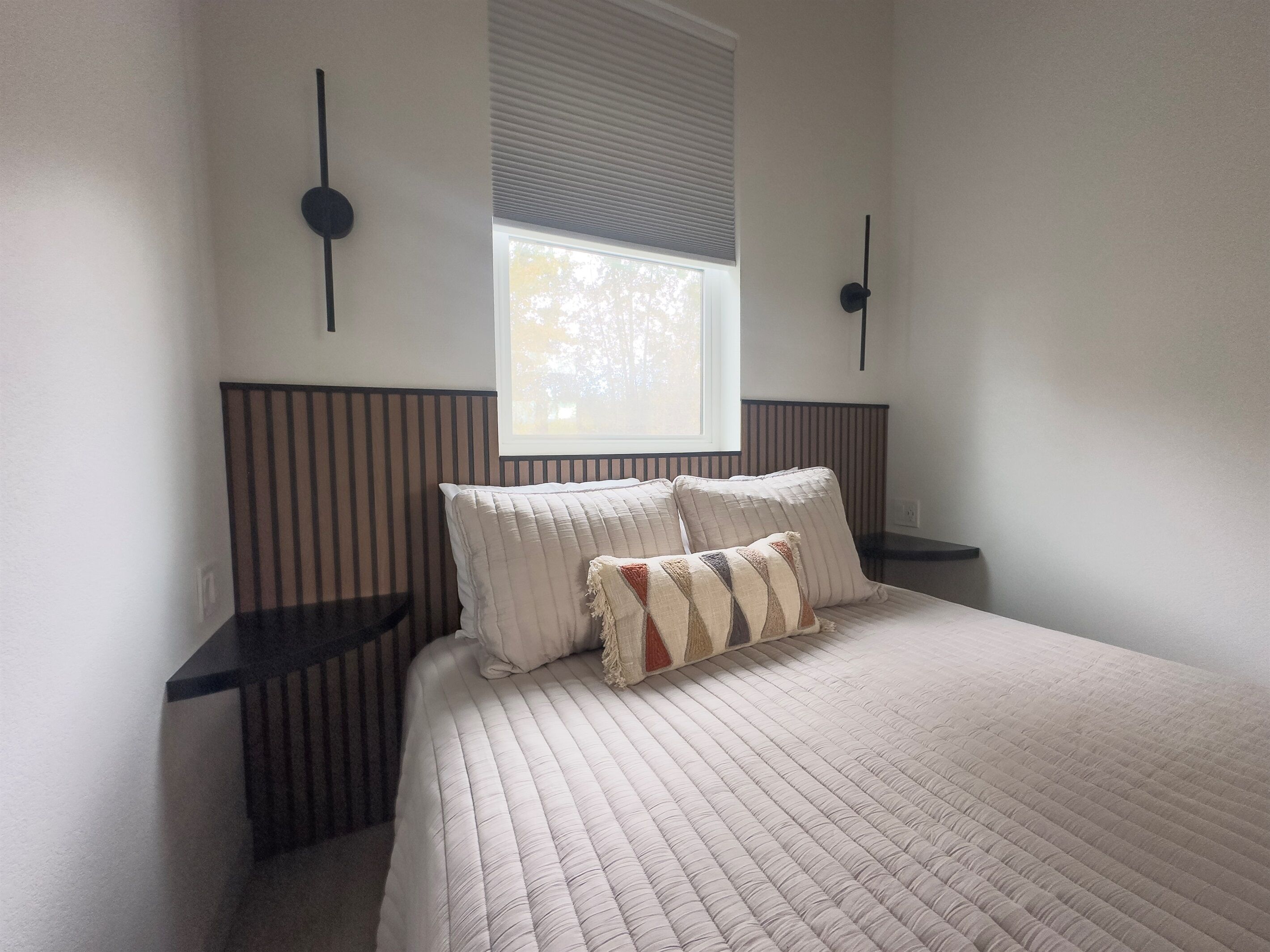A bedroom with a large bed, two bedside tables, a window with blinds, and wall-mounted light fixtures.