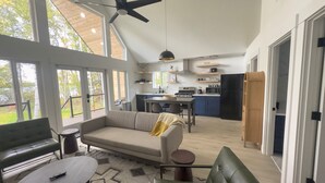 A spacious living area with a sofa, coffee table, and a dining table. A kitchen is visible in the background with modern appliances. - Lakefront Cottage with Sauna - #7 Dolly Varden (Wasilla)