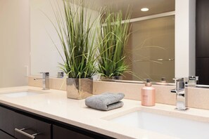 A bathroom with a double sink, a mirror, a plant, a towel, and a bottle.