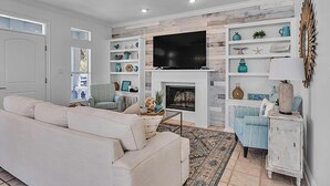 A living room with a white sofa, a fireplace, a TV mounted above it, and shelves with decorative items. - Jack Chasin' Birdies (Destin)