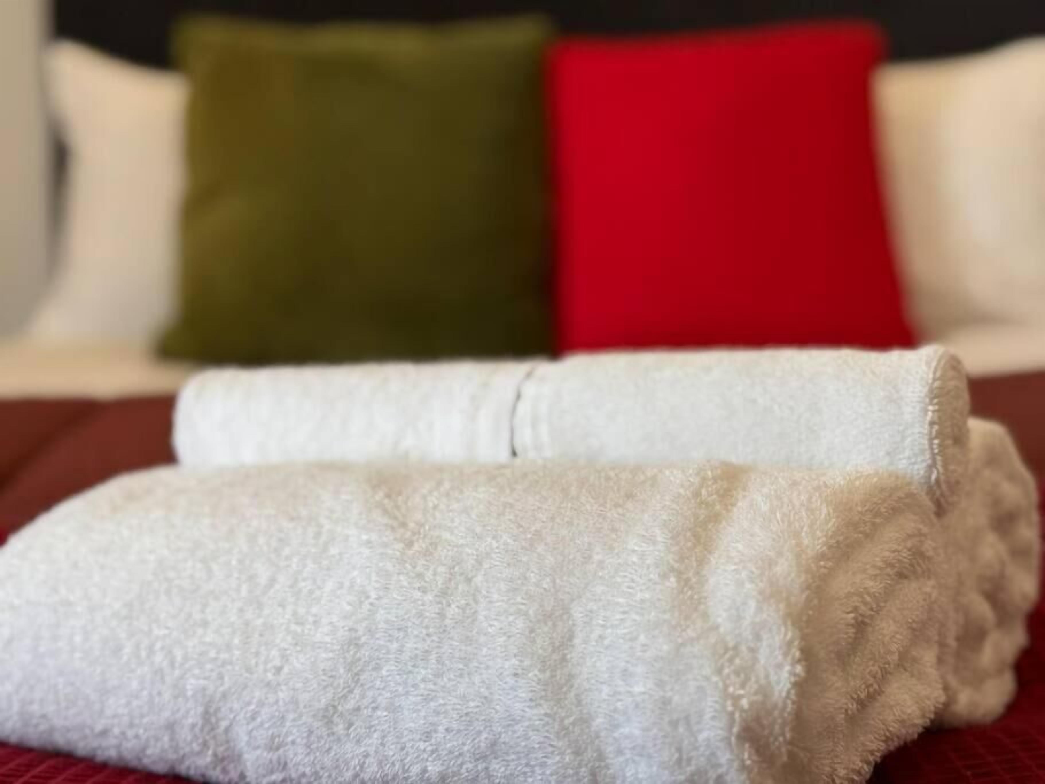 Een keurig opgevouwen witte handdoek op een bed, met een rood en groen kussen op de achtergrond.