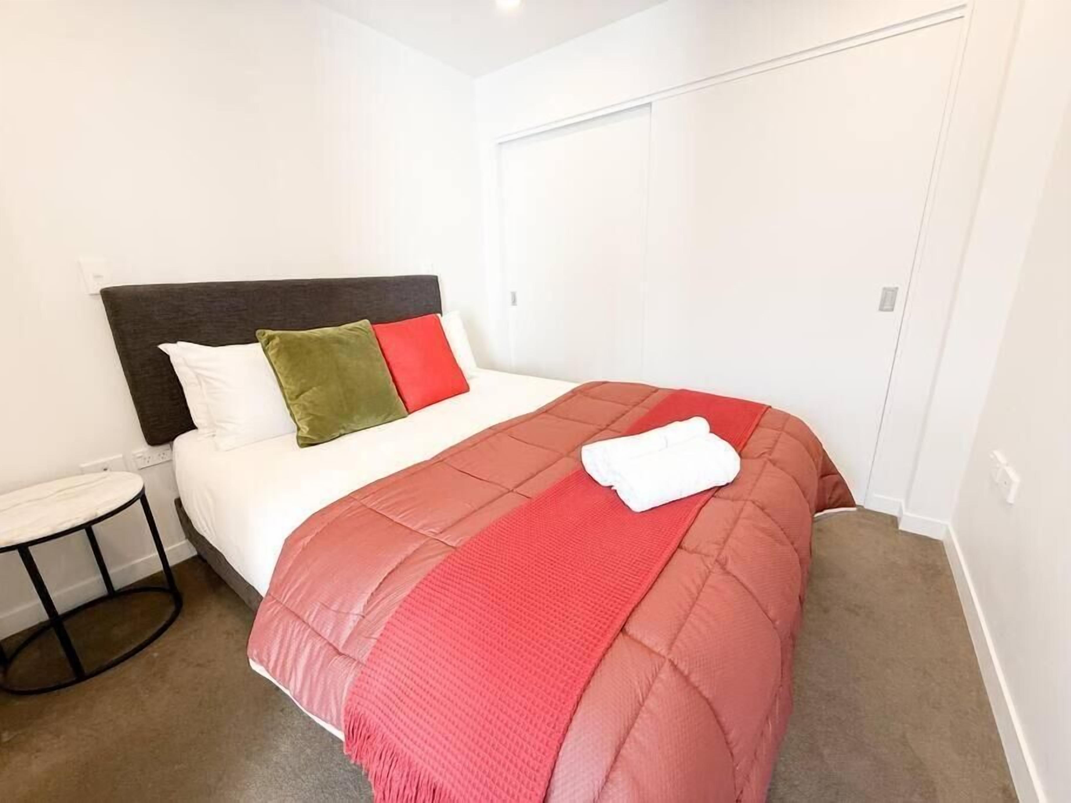Un lit bien fait, recouvert d’une couverture rouge et blanche, avec un oreiller vert et rouge et une serviette blanche posée sur le côté.
