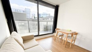 A modern living room with a beige sofa, a wooden dining table with two chairs, and a large window offering a view of city buildings. - Central Haven Studio on Queen Street  (Auckland)