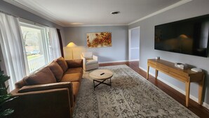 Ein Wohnzimmer mit braunem Sofa, einem weißen Sessel, einem runden Couchtisch, einem Fernseher auf einem Holzschrank und einem gemusterten Teppich.