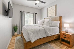 A bedroom with a bed, bedside table, lamp, ceiling fan, window with curtains, and a wall-mounted TV.