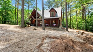 Une cabane en rondins dotée d’un porche et de fondations en pierre, entourée de grands arbres et surplombant un ciel dégagé.