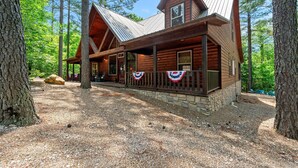 Une cabane en rondins, avec des fondations en pierre, une terrasse en bois et un toit en métal, entourée d’arbres.