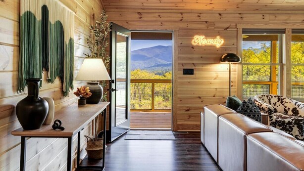 Interior de una cabaña de madera con sofá, una mesa con lámpara, un jarrón con flores y vistas a las montañas a través de amplios ventanales.