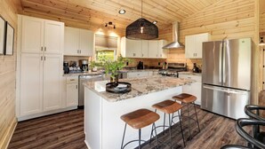 Cocina amplia con armarios de madera, una isla grande con taburetes, electrodomésticos de acero inoxidable y una lámpara colgante.