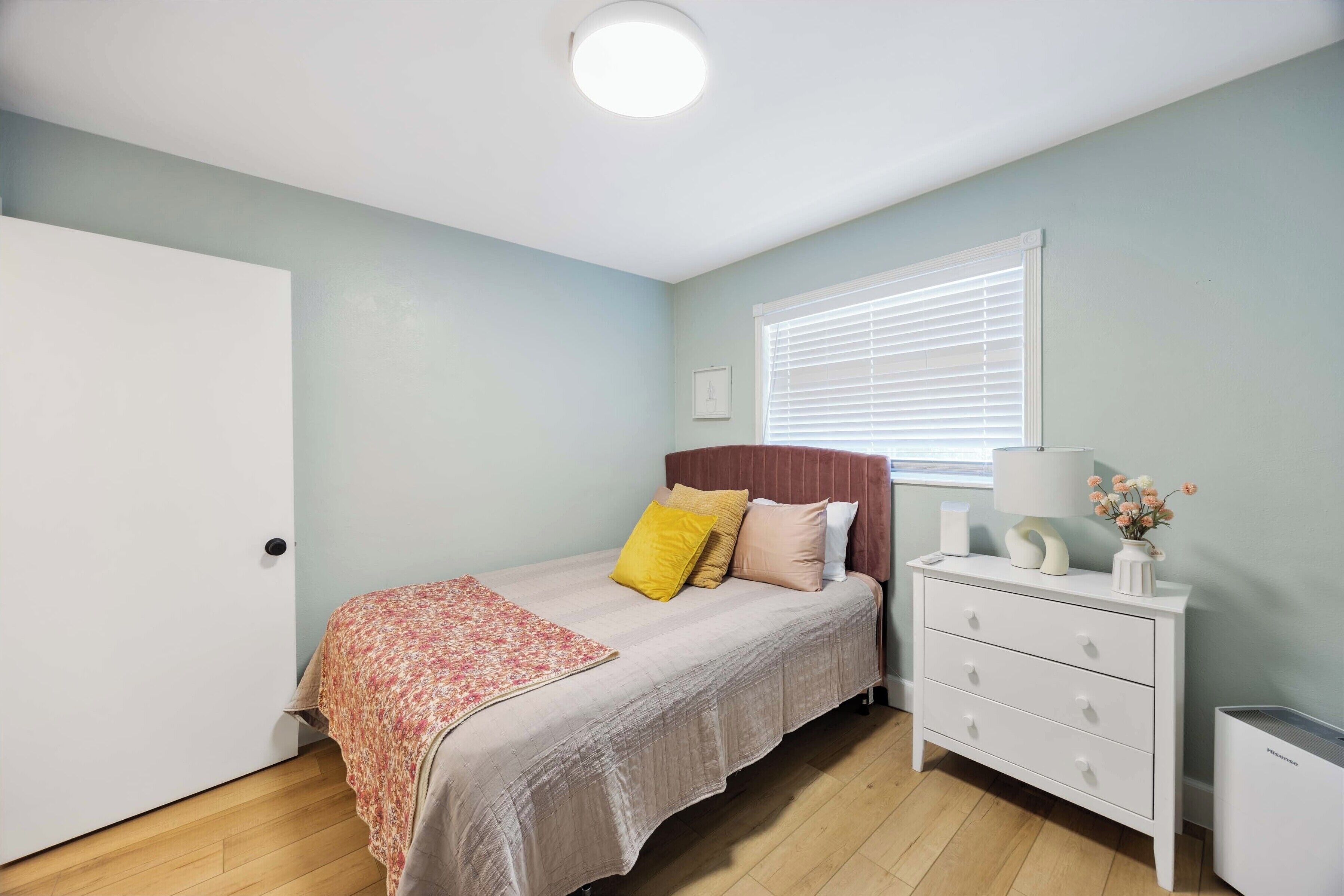 A bedroom with a bed, bedside table, and a window with blinds.