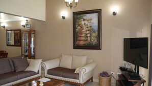 A living room with a beige sofa, a wooden coffee table, a painting of a staircase, and a flat-screen TV. - Beautiful Ocean View Home In San Antonio del Mar (Tijuana)