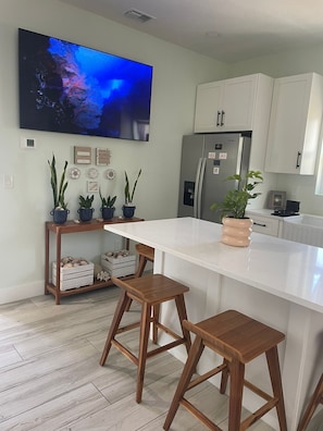 A modern kitchen with a large island, wooden stools, a refrigerator, and decorative plants. - New Coastal 2BR - Private Entrance - Near Beaches & Pinellas Trail - 2 Gig WiFi (Palm Harbor)