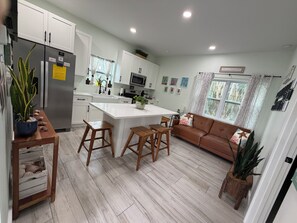A modern kitchen with white cabinets, a central island, wooden stools, a brown leather sofa, and a potted plant.