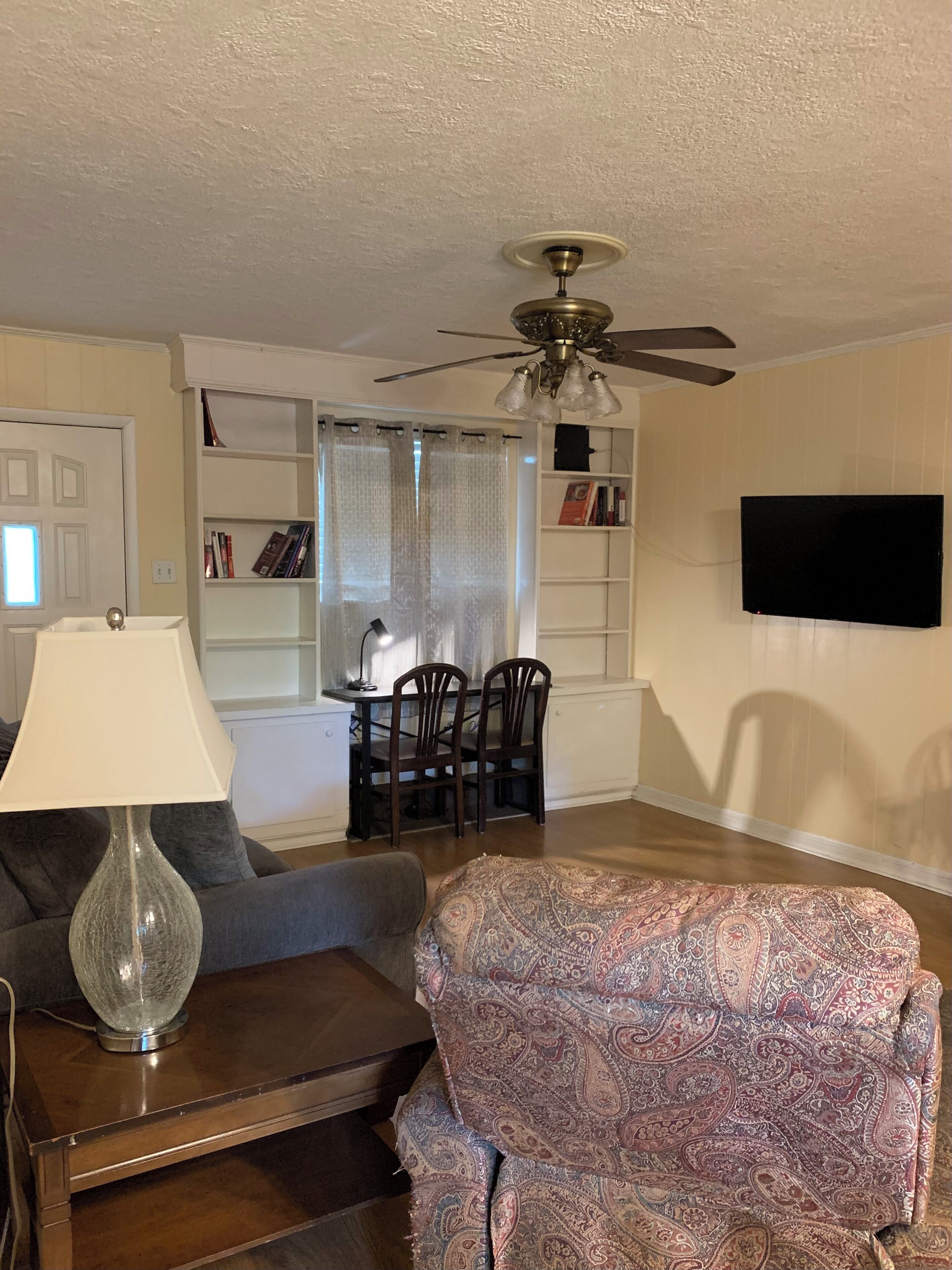 A living room with a ceiling fan, a TV mounted on the wall, a dining table with chairs, and a patterned rug.
