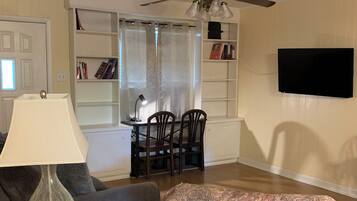 A living room with a ceiling fan, a TV mounted on the wall, a dining table with chairs, and a patterned rug.