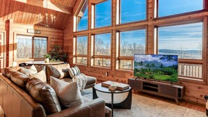 Interior acogedor de una cabaña con sofá de cuero, televisor de pantalla plana y ventanas amplias que ofrecen vistas panorámicas.