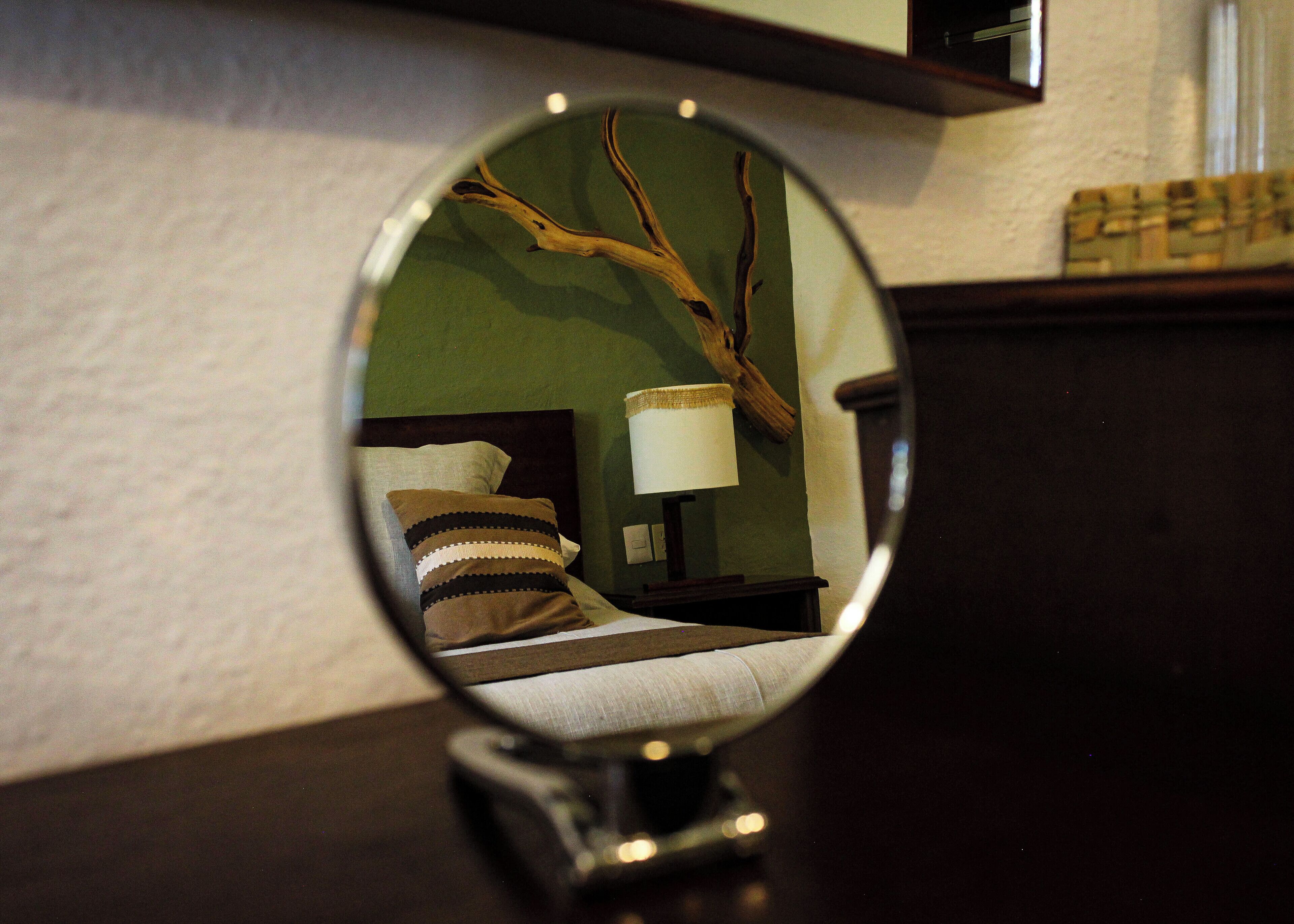 A round mirror reflecting a bed with a pillow, a bedside table with a lamp, and a wall with a textured pattern.