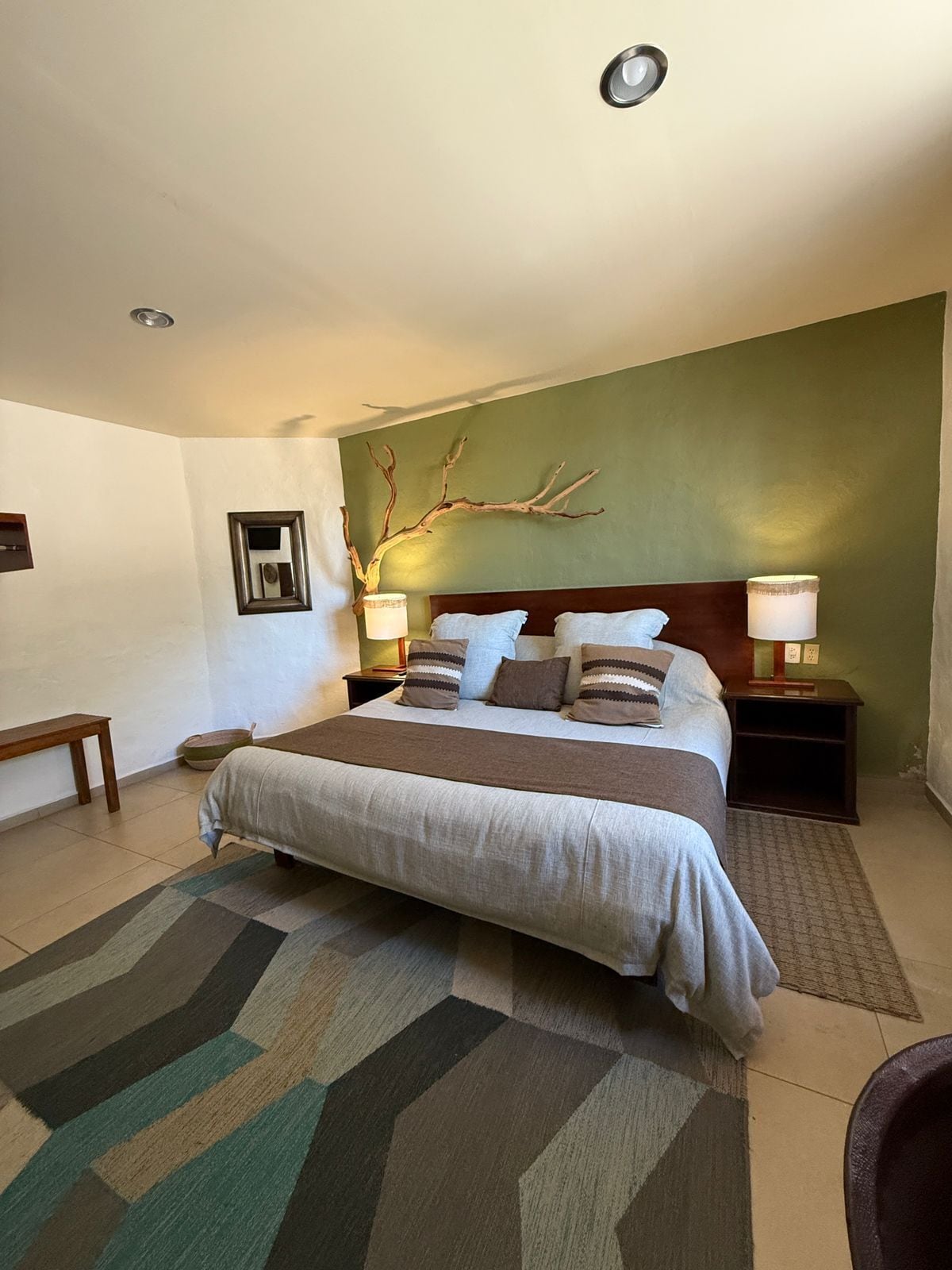 Une chambre à coucher bien aménagée, avec un grand lit, deux tables de chevet, une commode, un banc et une décoration murale en forme de branche.