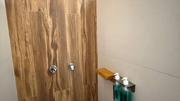 Baño con puerta de madera, zona de ducha y estante con artículos de aseo.