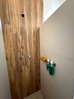 Baño con puerta de madera, zona de ducha y estante con artículos de aseo.