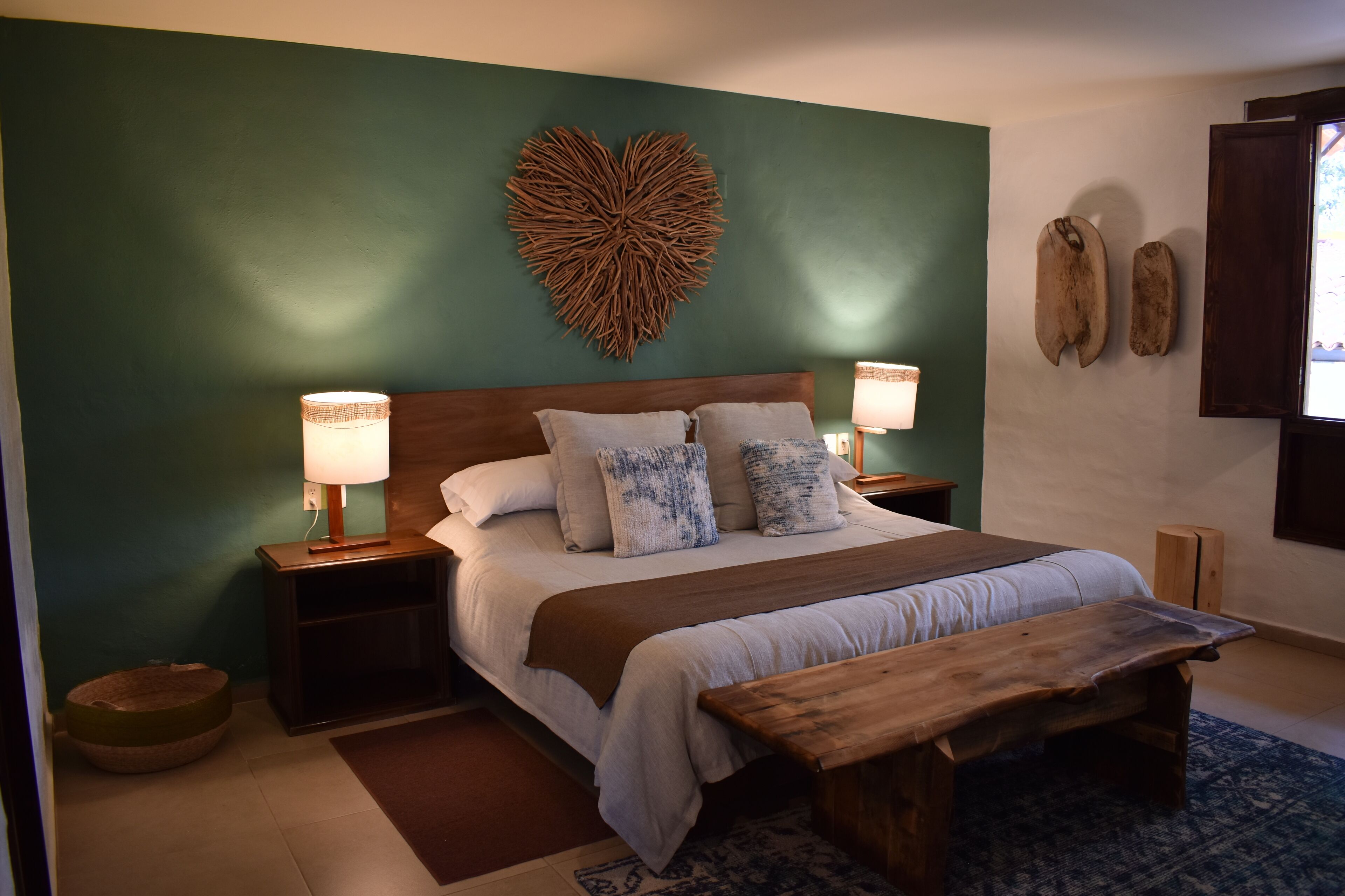 A bedroom with a large bed, two bedside lamps, a wooden bench, a heart-shaped wall decoration, and a small dog bed.