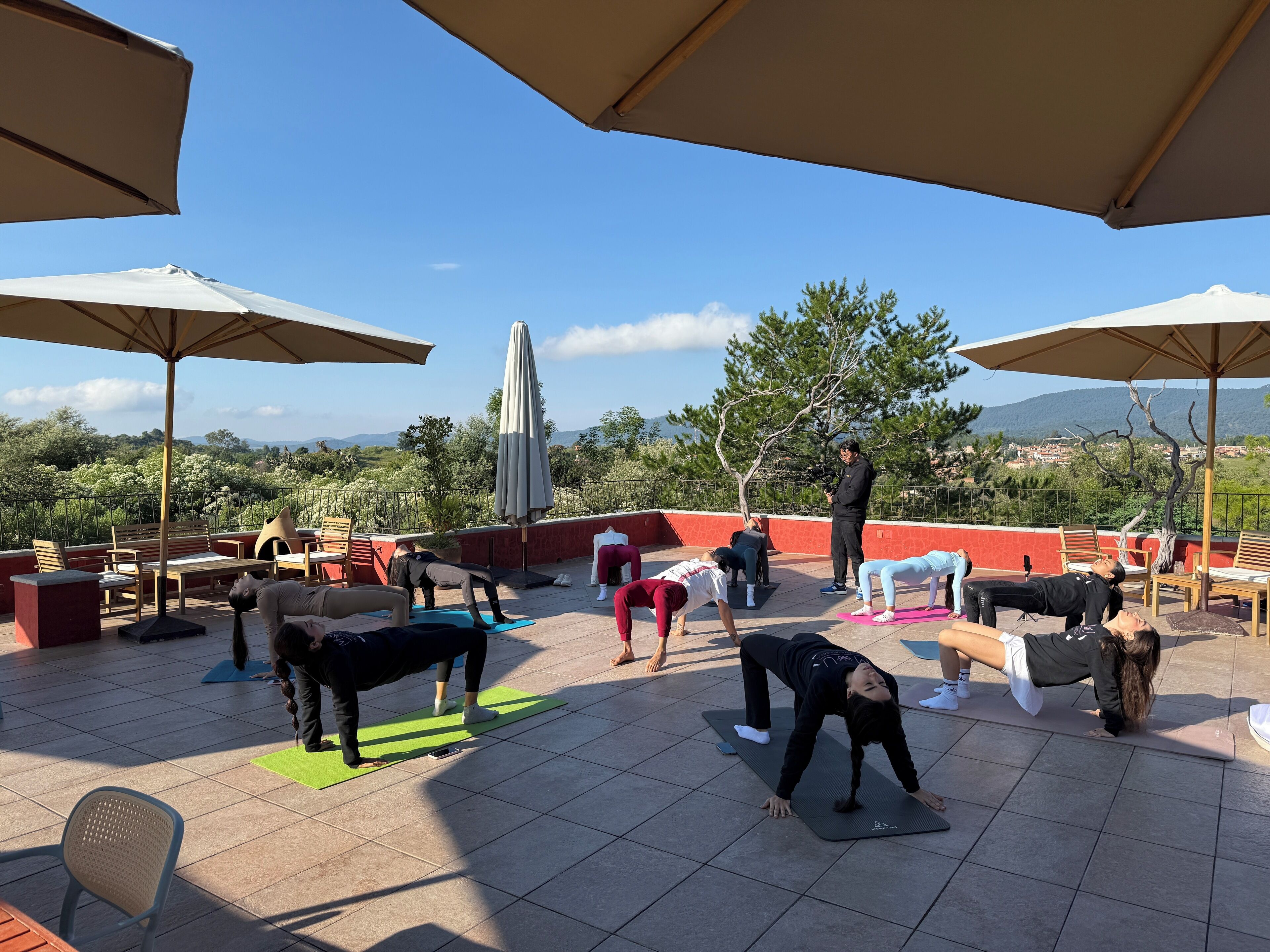 A rooftop yoga class with participants on mats, surrounded by outdoor furniture and umbrellas, overlooking a scenic landscape.