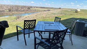 An outdoor patio with a table and chairs, overlooking a river and fields. - Cozy farmhouse in charming Great Falls with WiFi, AC and indoor parking (Great Falls)