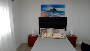 A bedroom with a bed, two nightstands, a painting of a beach scene, and a window with white curtains.