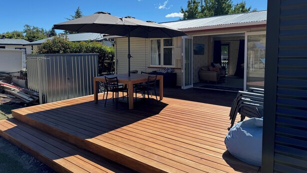 Eine Holzterrasse mit Tisch, Stühlen und einem Sonnenschirm, sowie eine Glasschiebetür zu einem Wohnbereich.