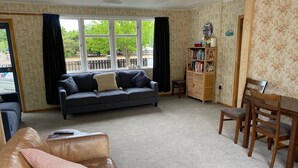 Ein Wohnzimmer mit einer Couch, einem Couchtisch mit Fernbedienungen, einem Holzregal und einem Esstisch mit Stühlen.