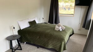 Ein ordentlich bezogenes Bett mit einer grünen Decke, ein kleiner Nachttisch mit Lampe und ein Fenster mit Vorhängen.