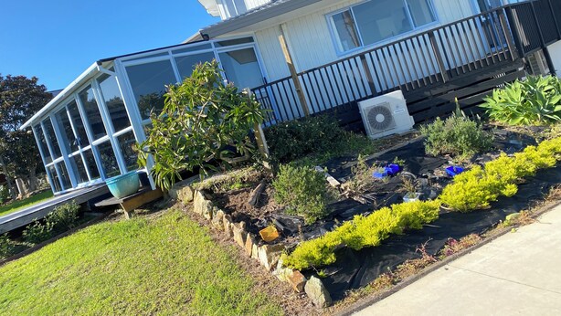 Una casa de dos pisos con un balcón, un ventanal grande y un jardín con diversas plantas.