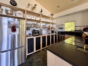 Une cuisine moderne équipée d’un réfrigérateur en acier inoxydable, d’étagères en bois où sont disposés des bocaux et des ustensiles de cuisine, d’une machine à café et d’une fenêtre donnant sur des arbres.