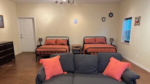 Een kamer met een grijze hoekbank, twee bedden met bijpassend beddengoed, een tafeltje en een raam met zonwering.
