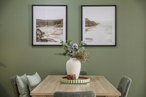 Un comedor con una mesa de madera, dos cuadros de paisajes enmarcados, un florero con follaje y sillas tapizadas de color gris.