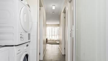 Un área de lavandería con lavadoras y secadoras apiladas, un pasillo que conduce a una habitación con cama y cortinas.