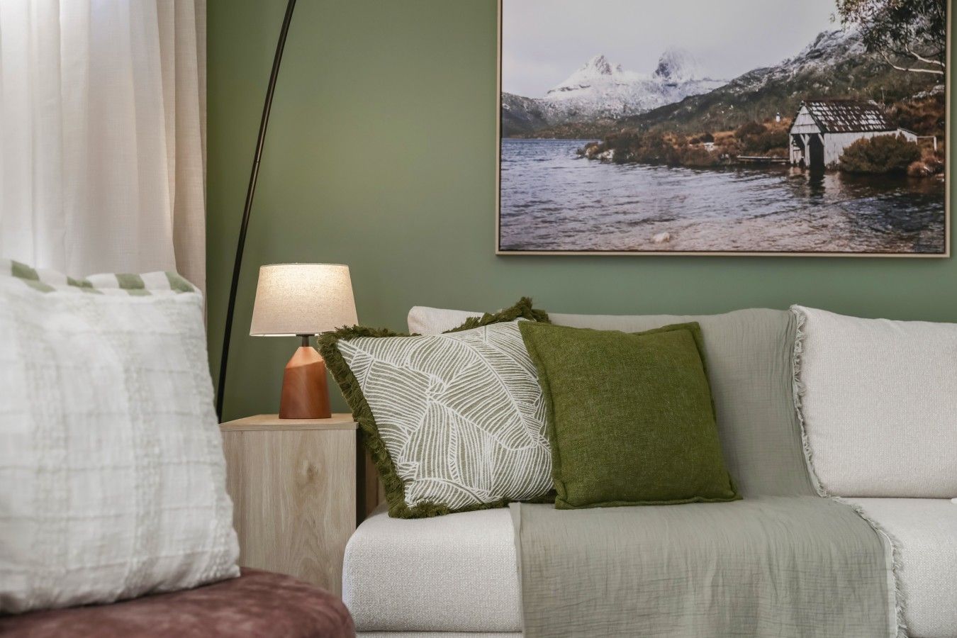 Una habitación con cama, una mesita de noche con lámpara, una pared verde y un cuadro enmarcado de un paisaje montañoso.