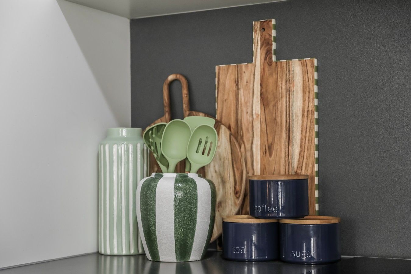 Un plan de travail de cuisine avec une planche à découper en bois, un vase à rayures vertes et blanches, un ensemble de cuillères vertes et trois récipients bleu foncé étiquetés « café », « thé » et « sucre ».