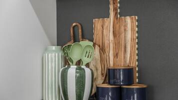 Un plan de travail de cuisine avec une planche à découper en bois, un vase à rayures vertes et blanches, un ensemble de cuillères vertes et trois récipients bleu foncé étiquetés « café », « thé » et « sucre ».