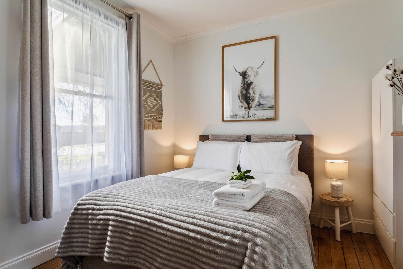 A hotel room with a bed, bedside lamps, a window with curtains, a wall-mounted artwork, and a small stool.