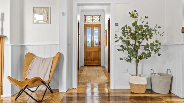 A hallway with a wooden floor, a chair, a potted plant, and a door leading to another room.