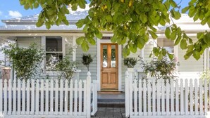 A white picket-fenced house with a wooden front door and two potted plants on either side.