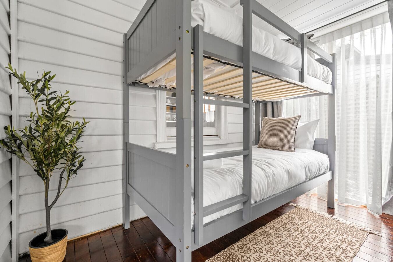 Ein Stockbett mit weißer Bettwäsche, ein Blumentopf und ein gemustertes Teppich in einem Zimmer mit Holzboden und weißen Wänden.