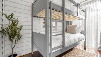 A bunk bed with white bedding, a potted plant, and a patterned rug in a room with wooden flooring and white walls.